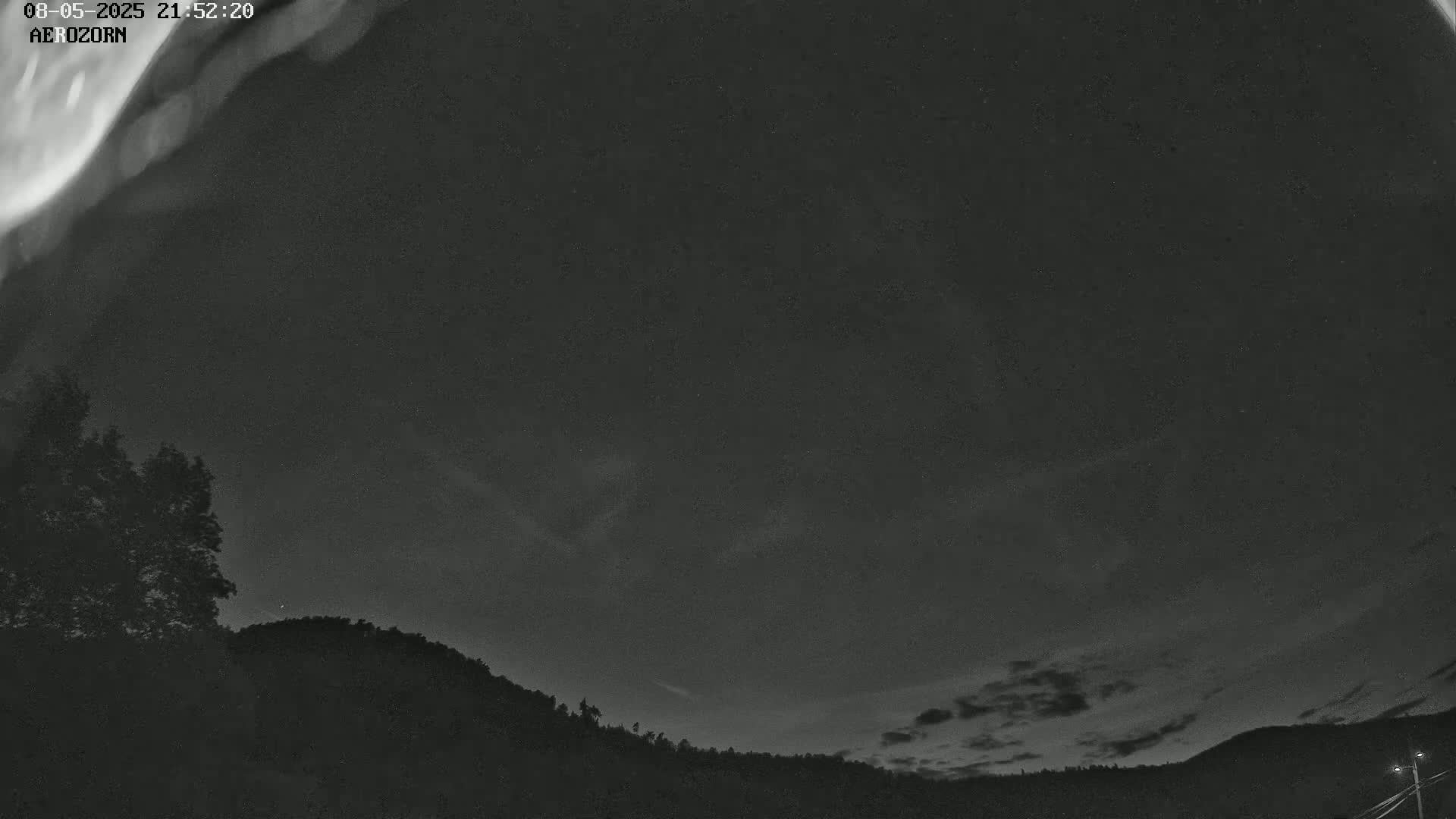 A nighttime, low-light image shows a dark sky with faint clouds and what appears to be a faint aurora borealis over a silhouetted landscape of trees and hills.