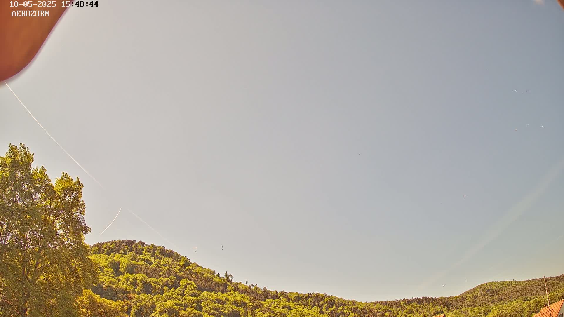 A mostly clear, pale sky over a lush green hill dotted with several small, distant objects possibly birds or aircraft.