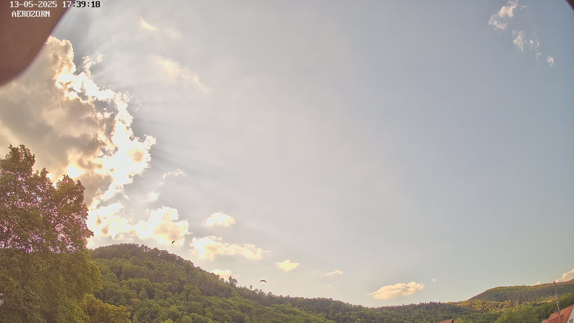 A paraglider flies in a partly cloudy sky above a lush green hillside.