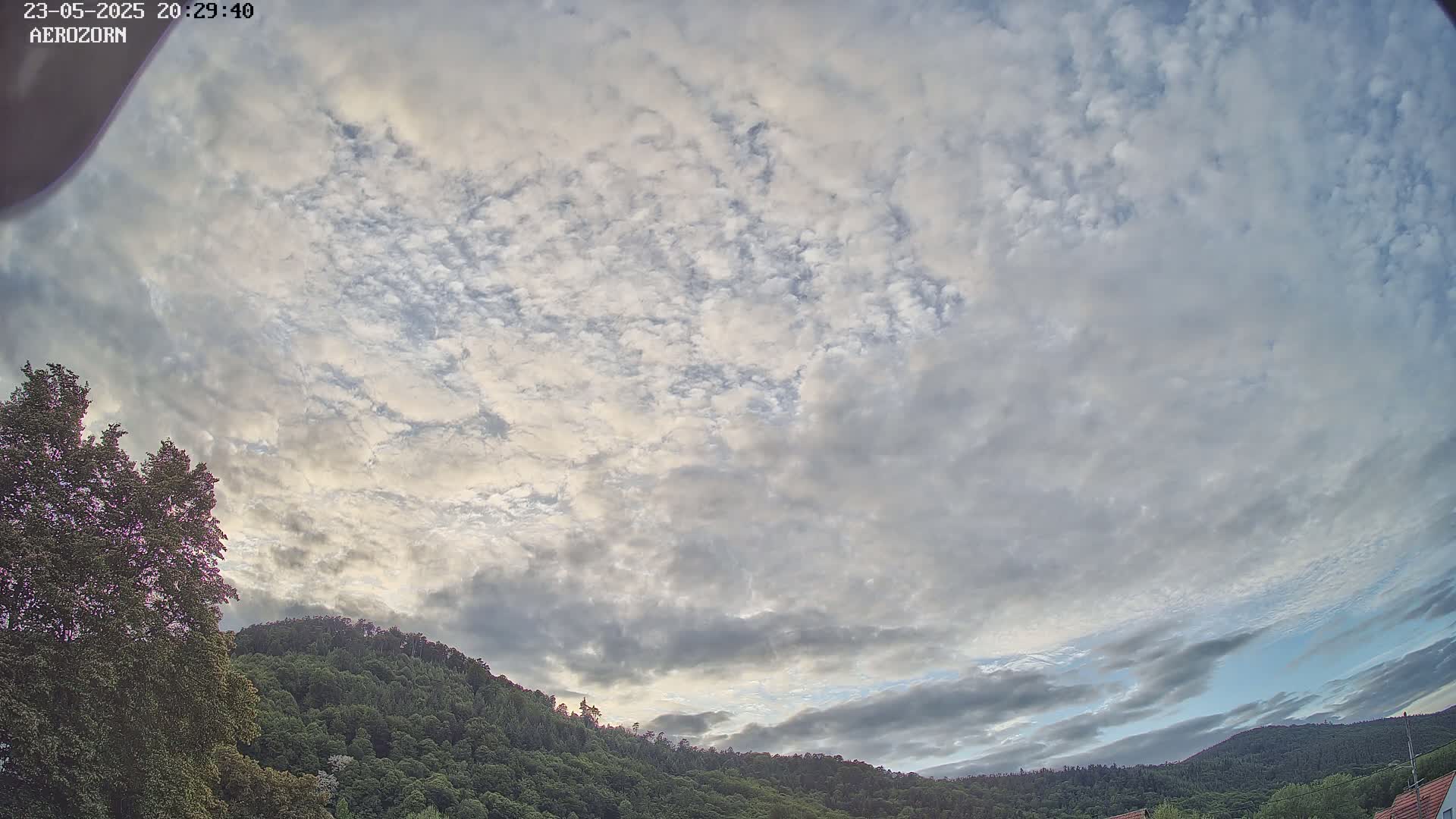 A mostly cloudy sky with numerous small, fluffy clouds hangs above a line of dark green forested hills.