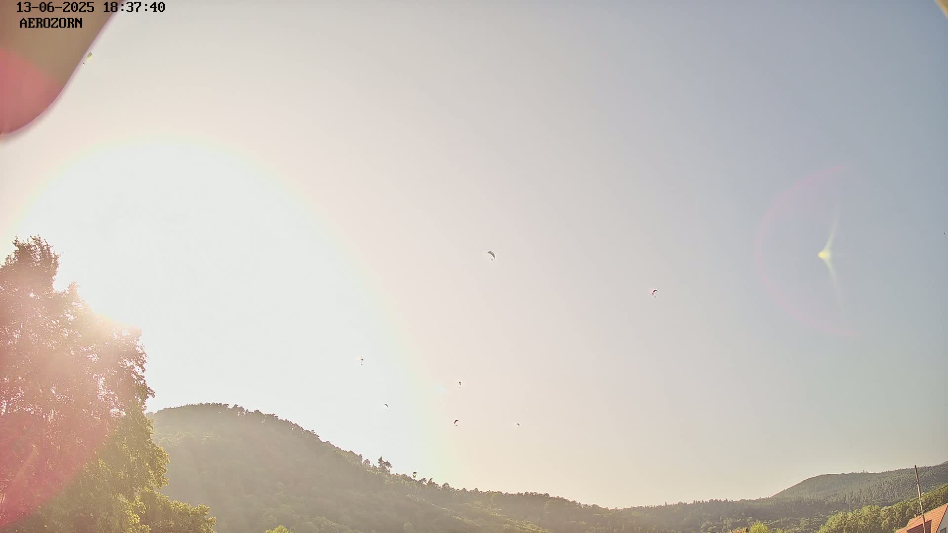 Several paragliders fly in a hazy, bright sky above a line of green hills, with the sun shining brightly in the lower left corner.