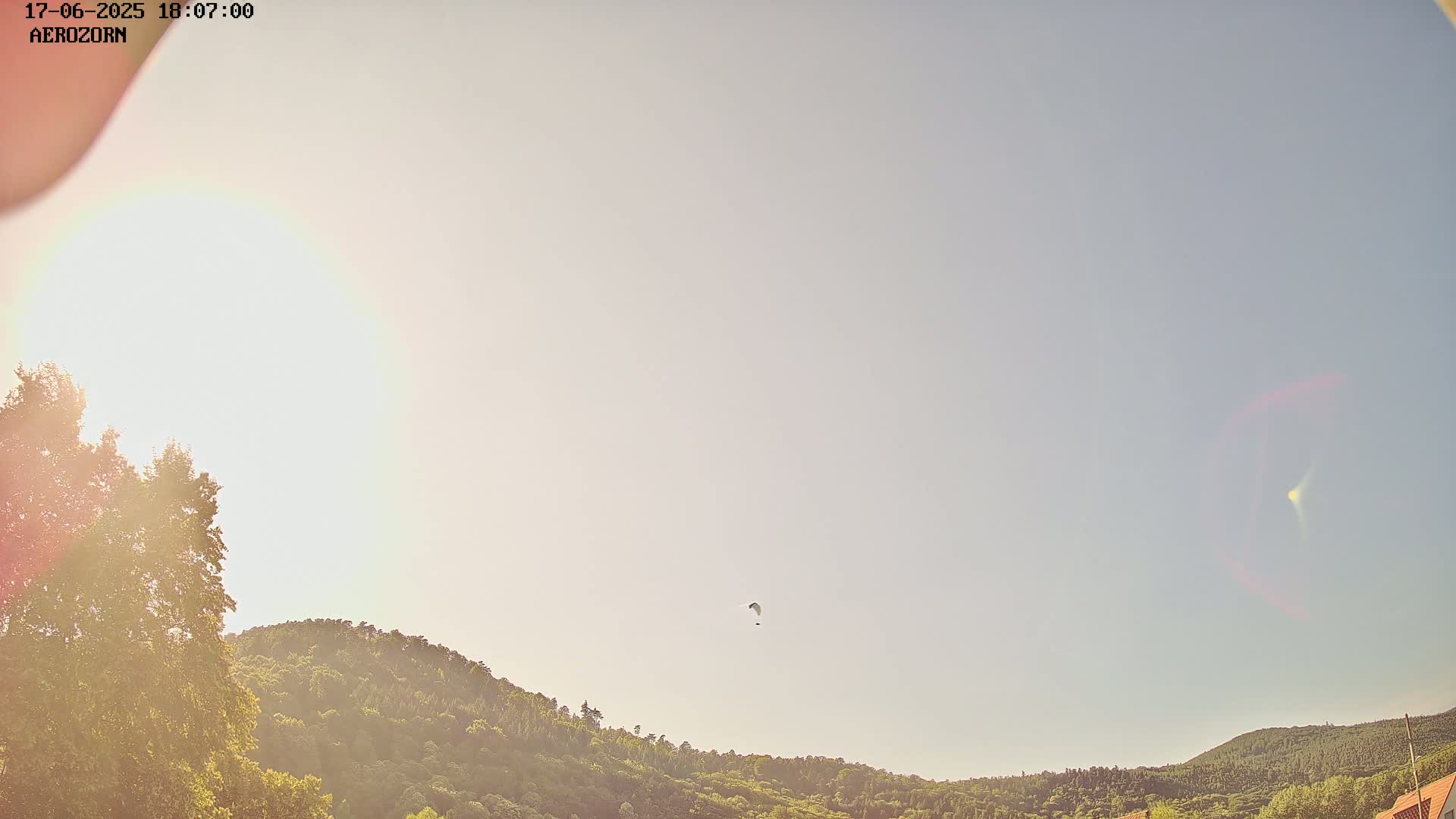 A paraglider is seen flying in a mostly clear sky above a line of green hills on a sunny day.