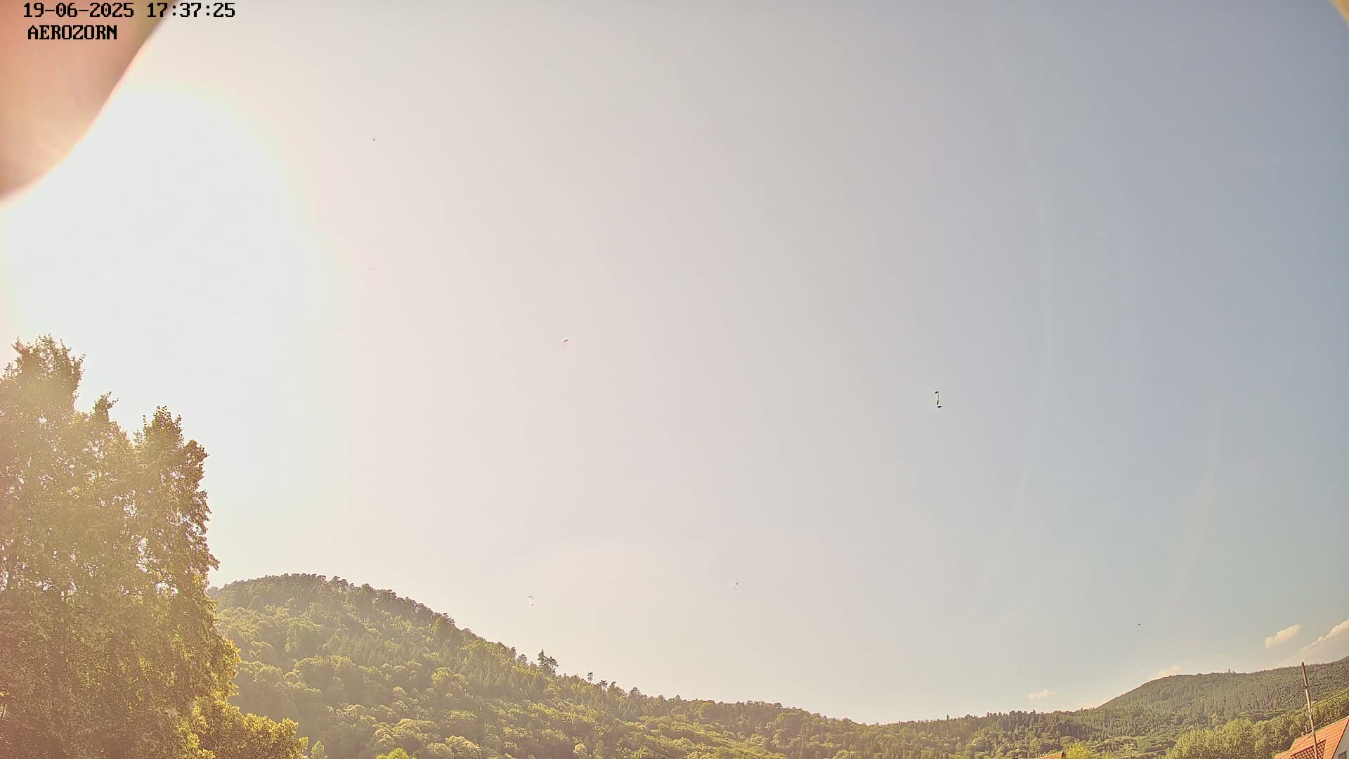 A bright, mostly clear sky is visible above a line of green hills and trees, with several small objects, possibly paragliders, flying in the distance.