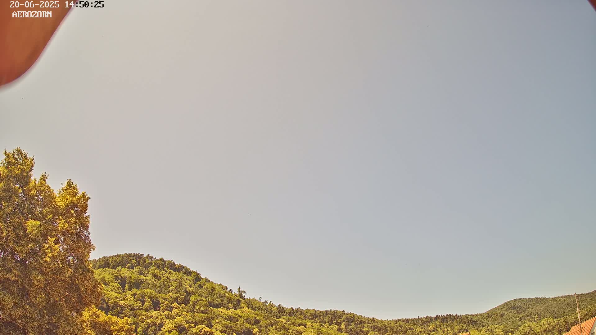 Under a mostly clear, light-grey sky, green-leafed hills and trees are visible.