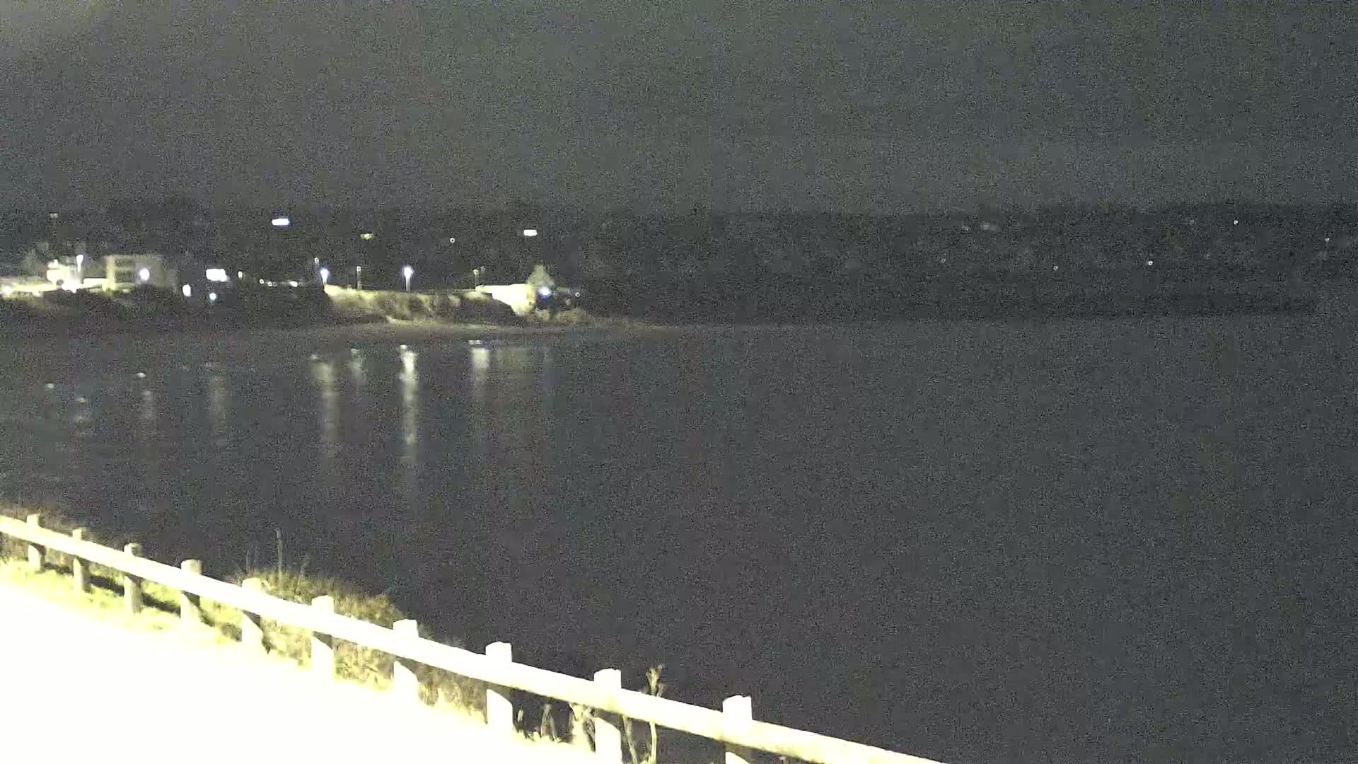 A nighttime view shows a calm body of water reflecting lights from a distant shoreline, with a wooden fence and road visible in the foreground under dark skies.