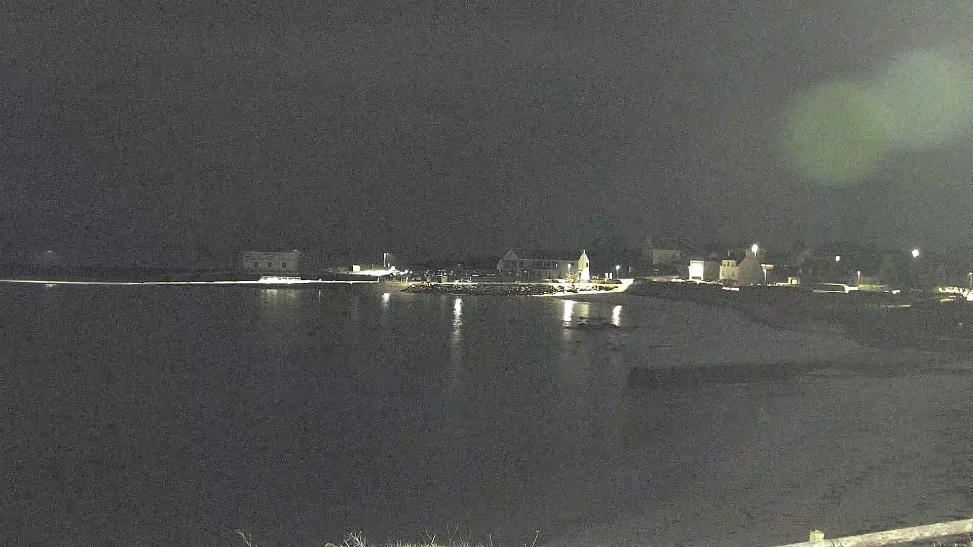 A nighttime view of a calm body of water reflecting the lights of buildings along a shoreline under a dark, clear sky.
