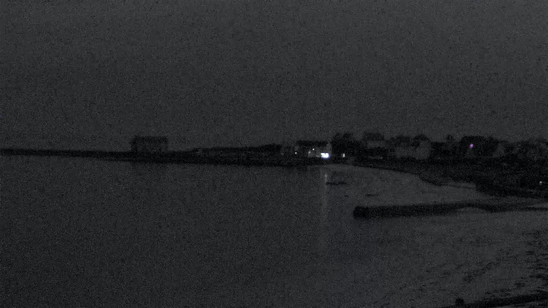 A dark, nighttime view shows a calm body of water reflecting a dimly lit shoreline with buildings and trees.