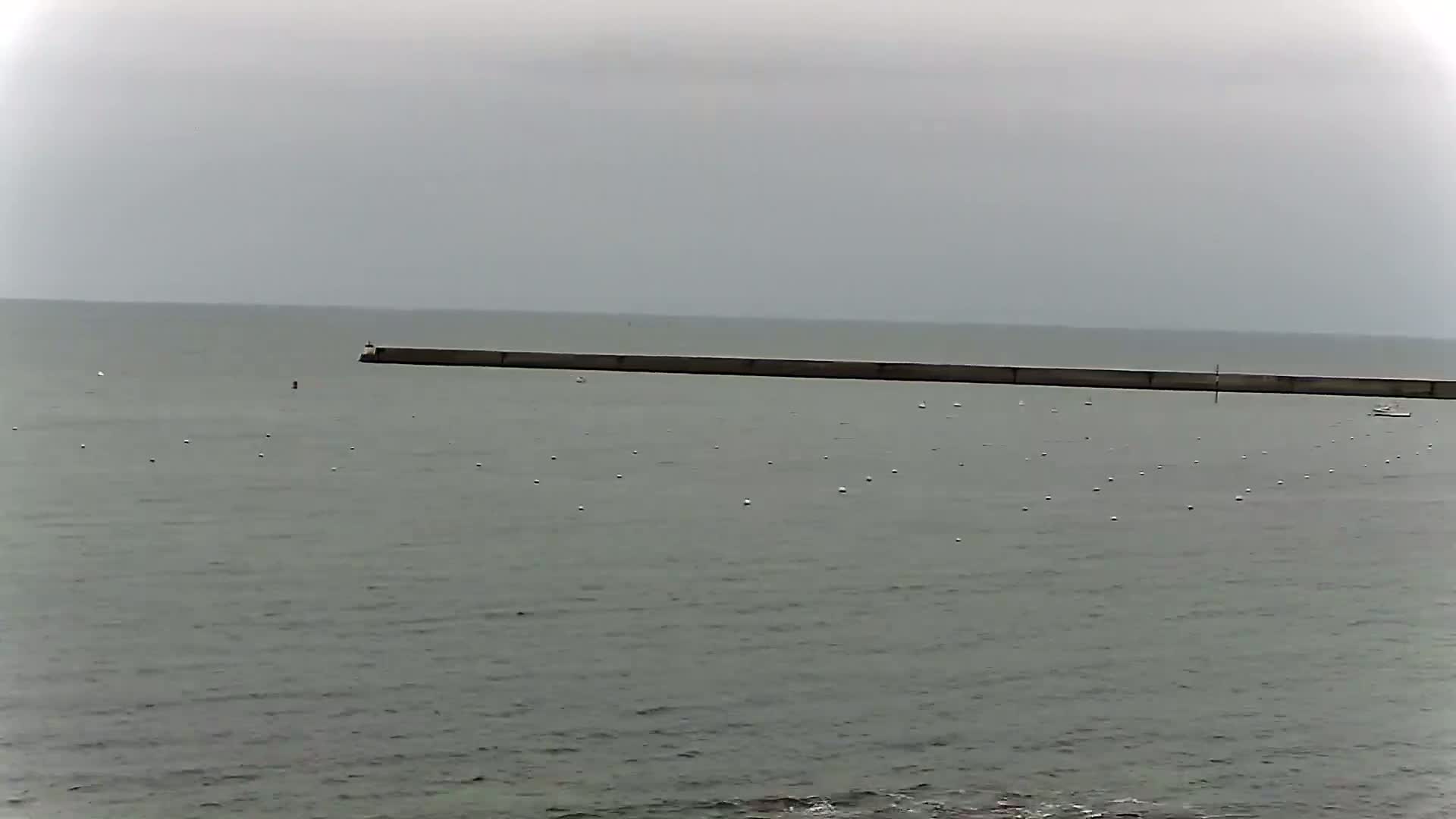 A long, low breakwater stretches across a calm, gray sea under an overcast sky, with several small white buoys scattered in the water.