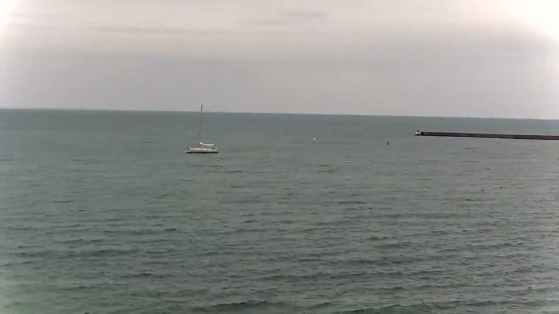 A sailboat floats on a calm, gray sea under an overcast sky, with a long breakwater visible in the distance.