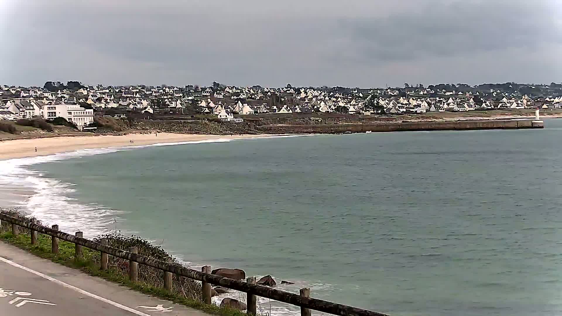 A sandy beach with small waves laps a calm, greenish-grey sea under an overcast sky, next to a town of white houses on a low cliff.