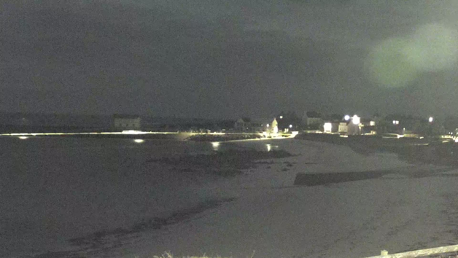 A dark nighttime scene shows a calm body of water next to a shoreline with sparsely lit buildings in the background under a cloudy sky.