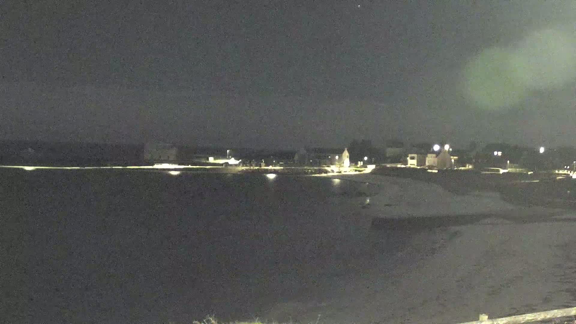 A nighttime view of a calm body of water reflecting the lights of a shoreline development under a dark, clear sky.
