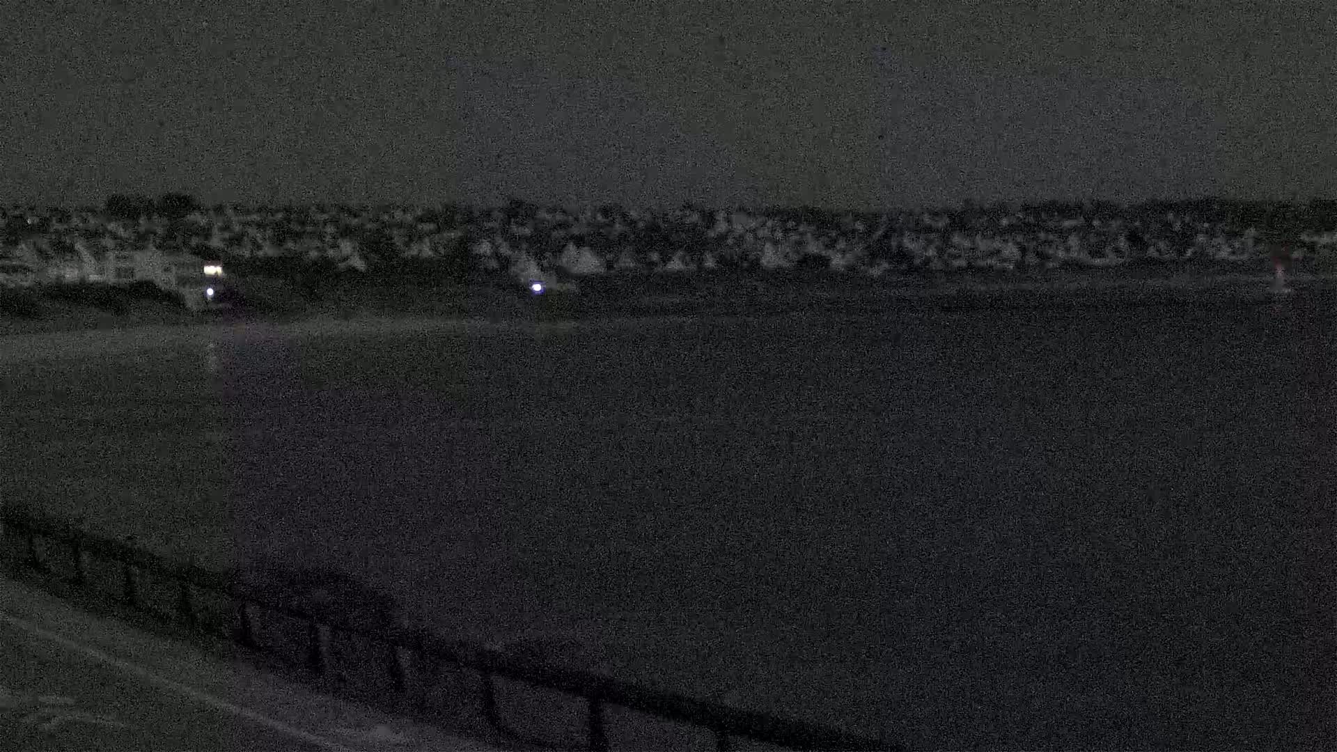 At night, a dark body of water is in the foreground, with a dark shoreline behind it containing a few small lights.