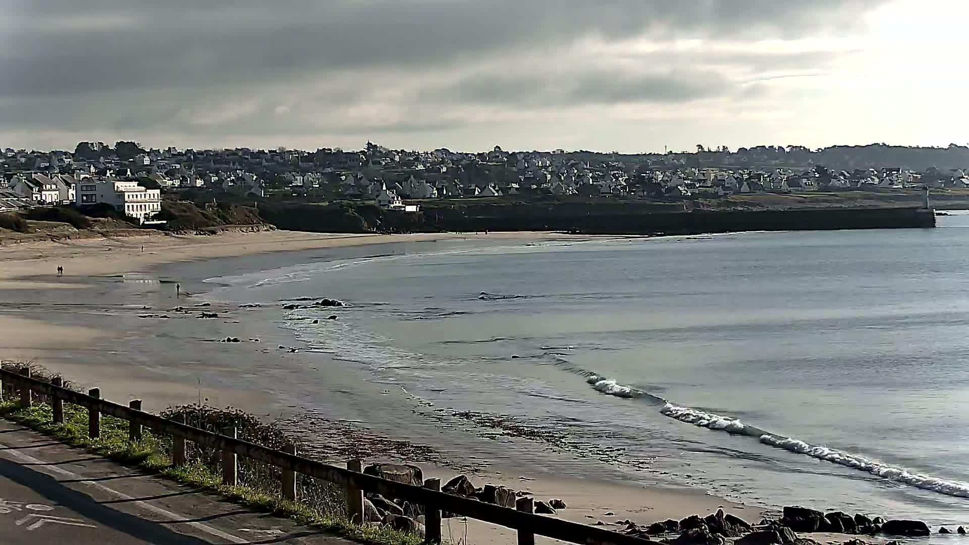 A sandy beach with gentle waves under a cloudy sky, overlooks a town nestled on a cliff.