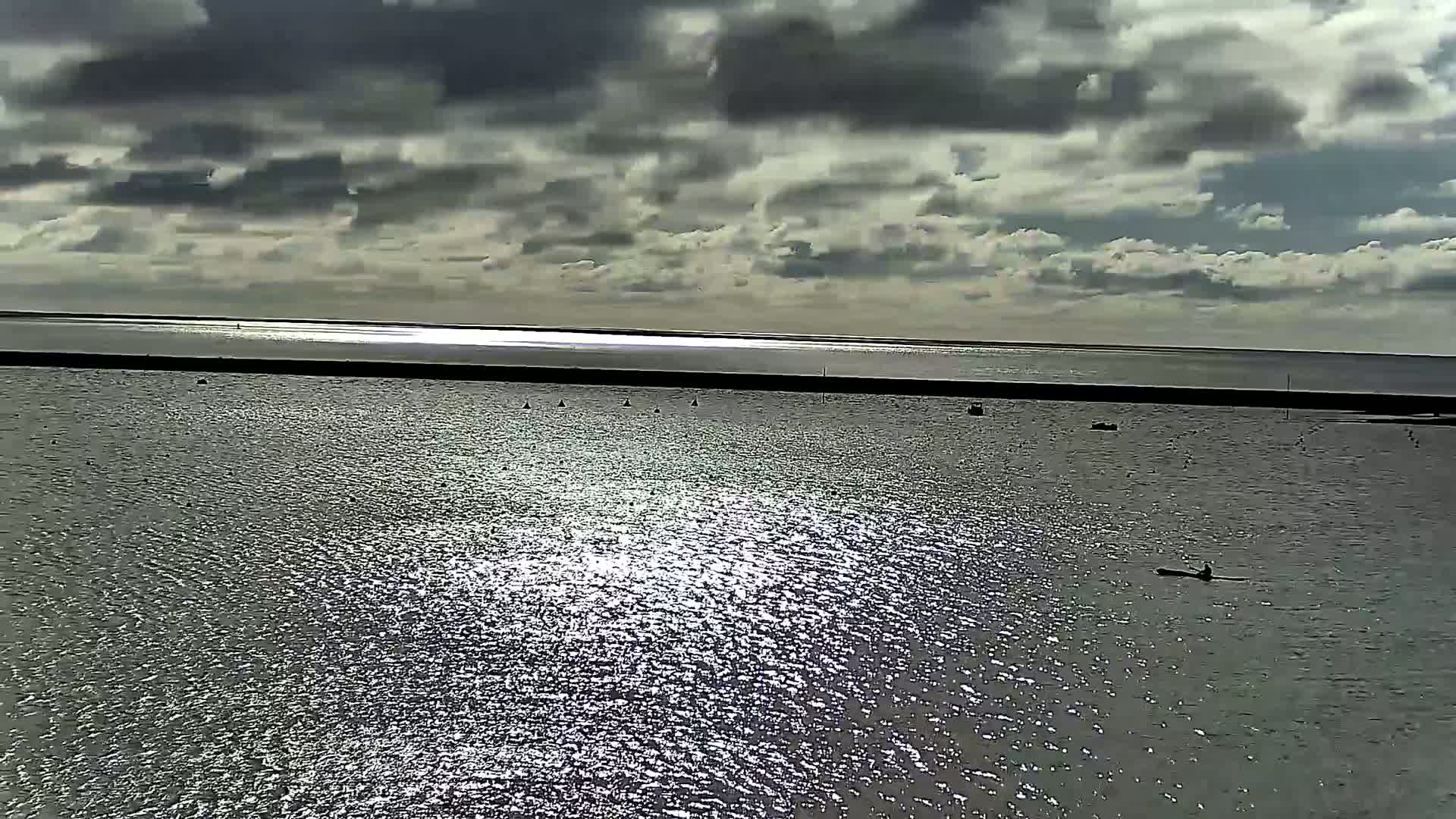 A mostly cloudy day reveals a calm body of water reflecting sunlight, with several small boats and a kayaker visible.