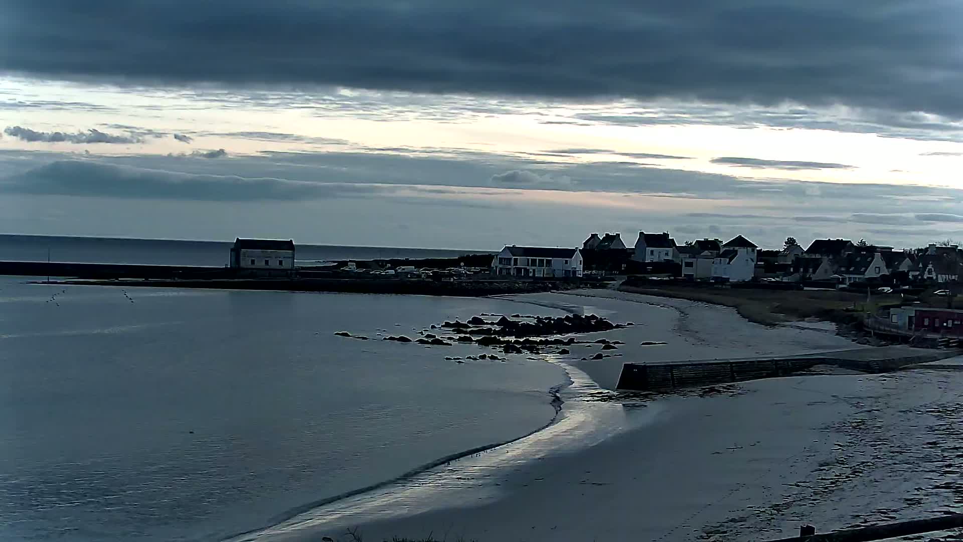 A coastal town with a calm, overcast sky sits beside a tranquil body of water, a sandy beach, and a rocky shoreline.