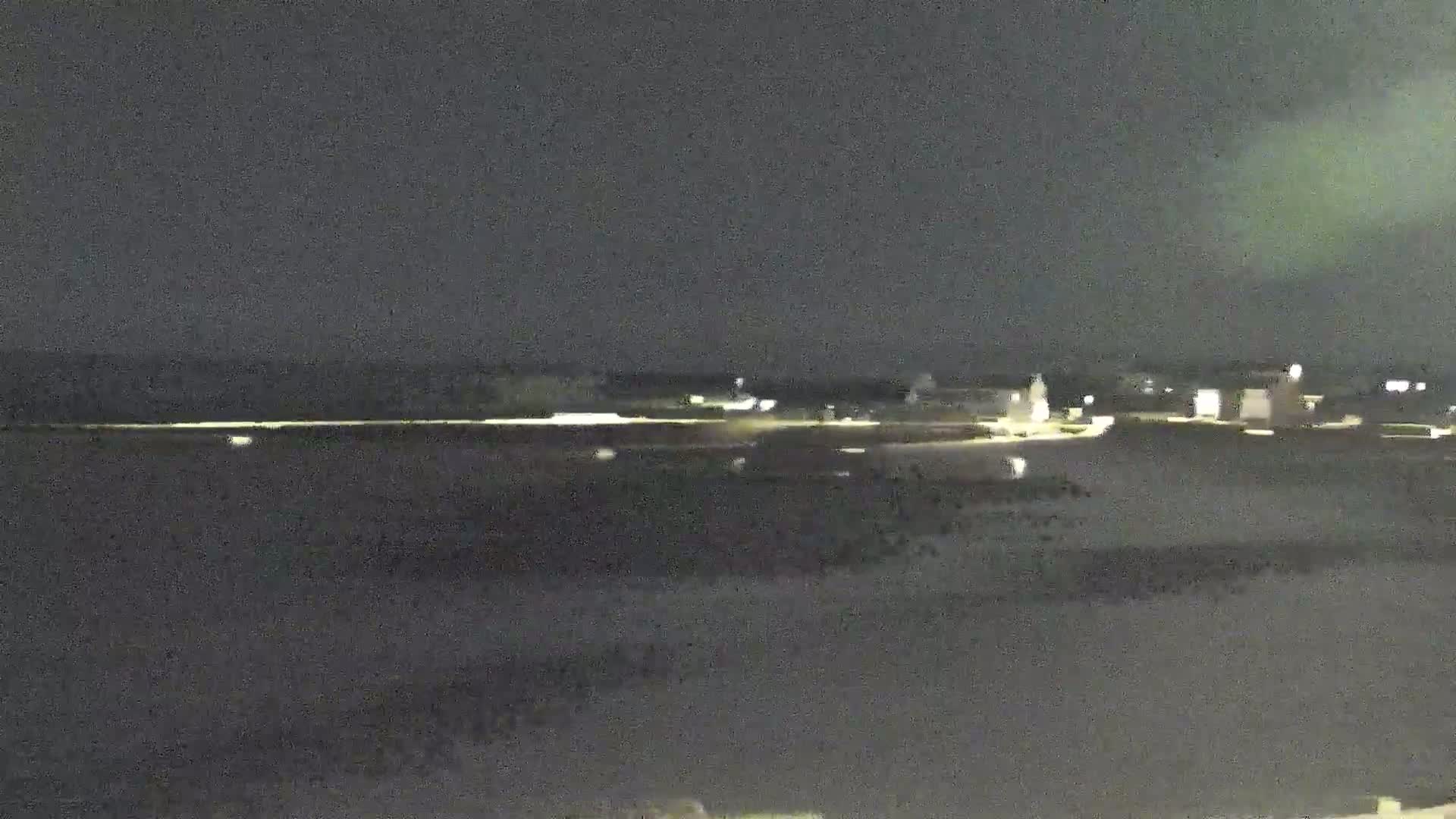 A nighttime, long shot shows a dimly lit coastal town across a dark body of water under a cloudy sky.