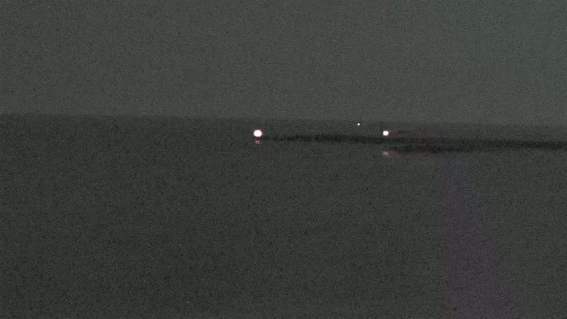 At night, a dark body of water reflects a few faint lights from a distant shoreline under a dark sky.