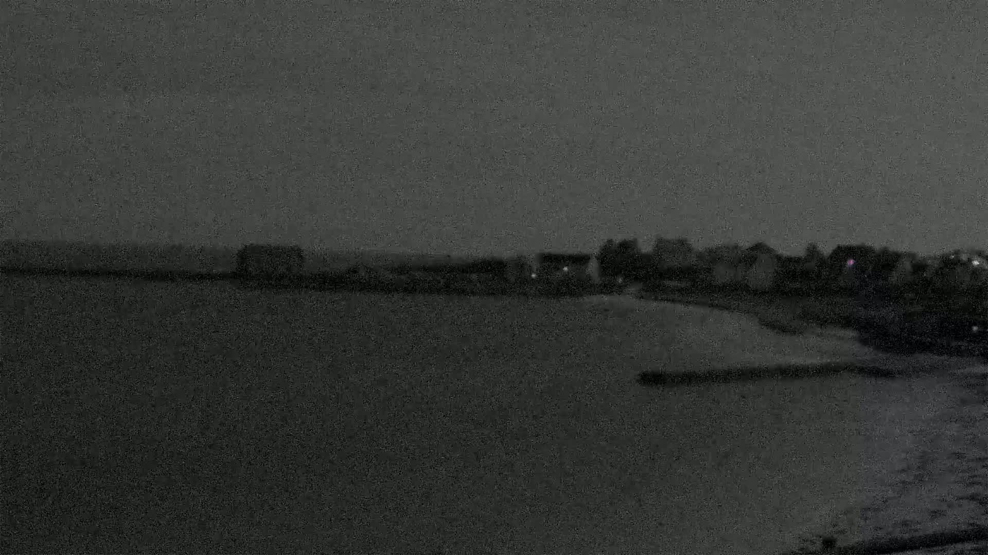 A dark, nighttime coastal scene shows a body of water in the foreground and a shoreline with buildings under a gray sky.