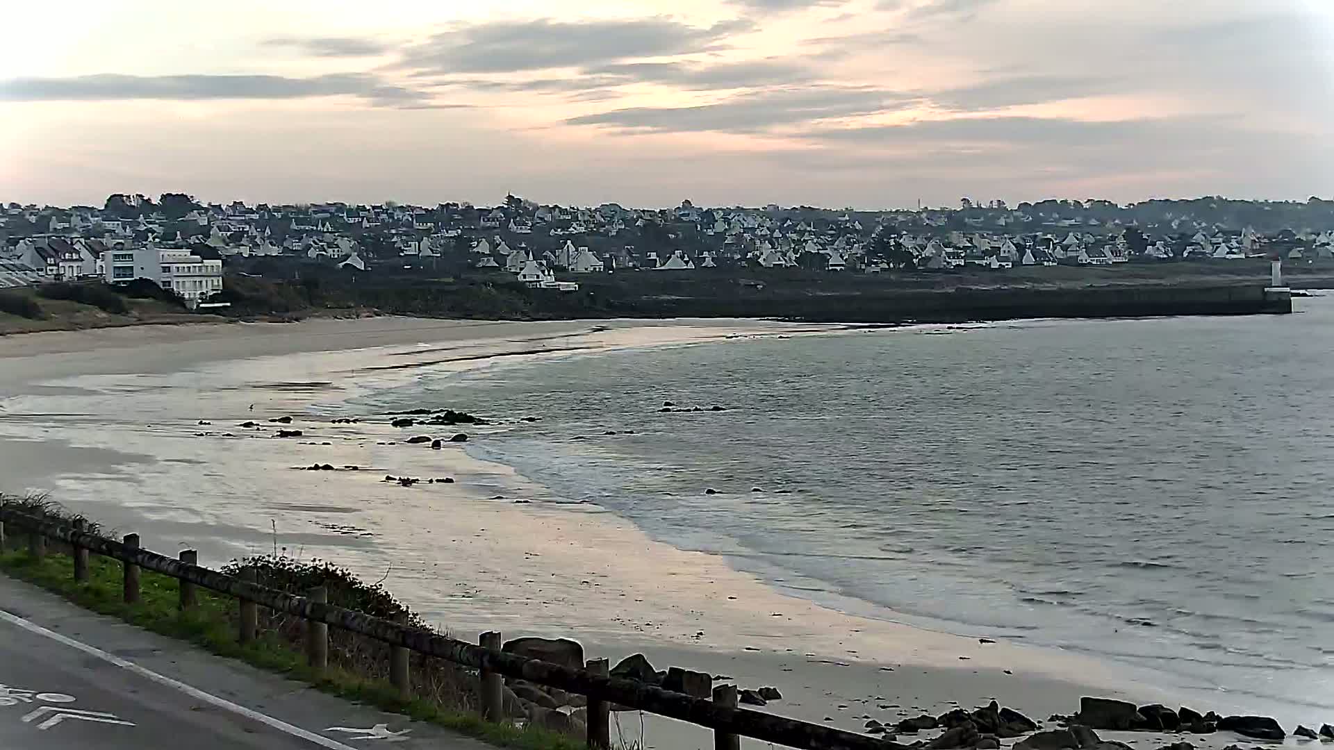 A sandy beach with gentle waves curves around a coastline, under a cloudy, dusky sky, towards a town of white houses.