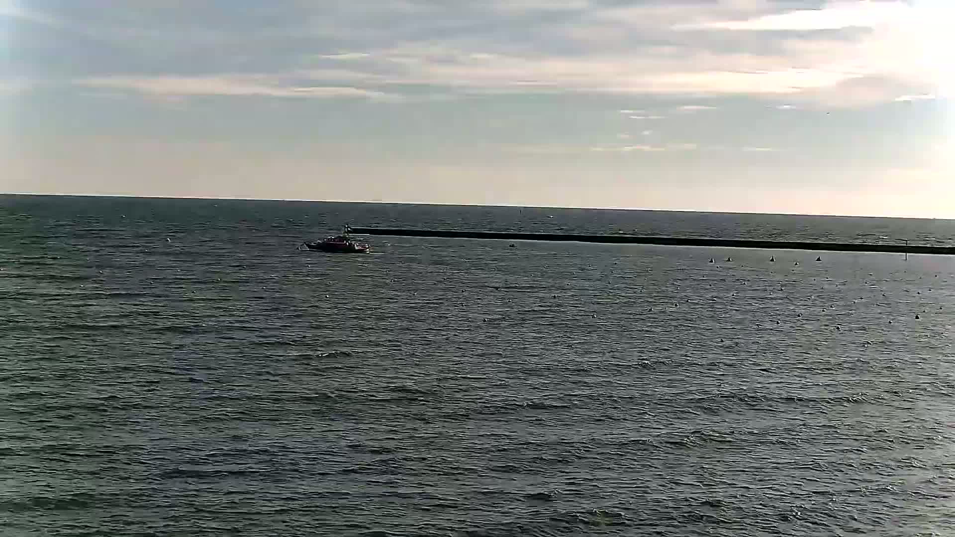 A small boat is on a dark-grey sea under a partly cloudy sky, near a long, low breakwater.