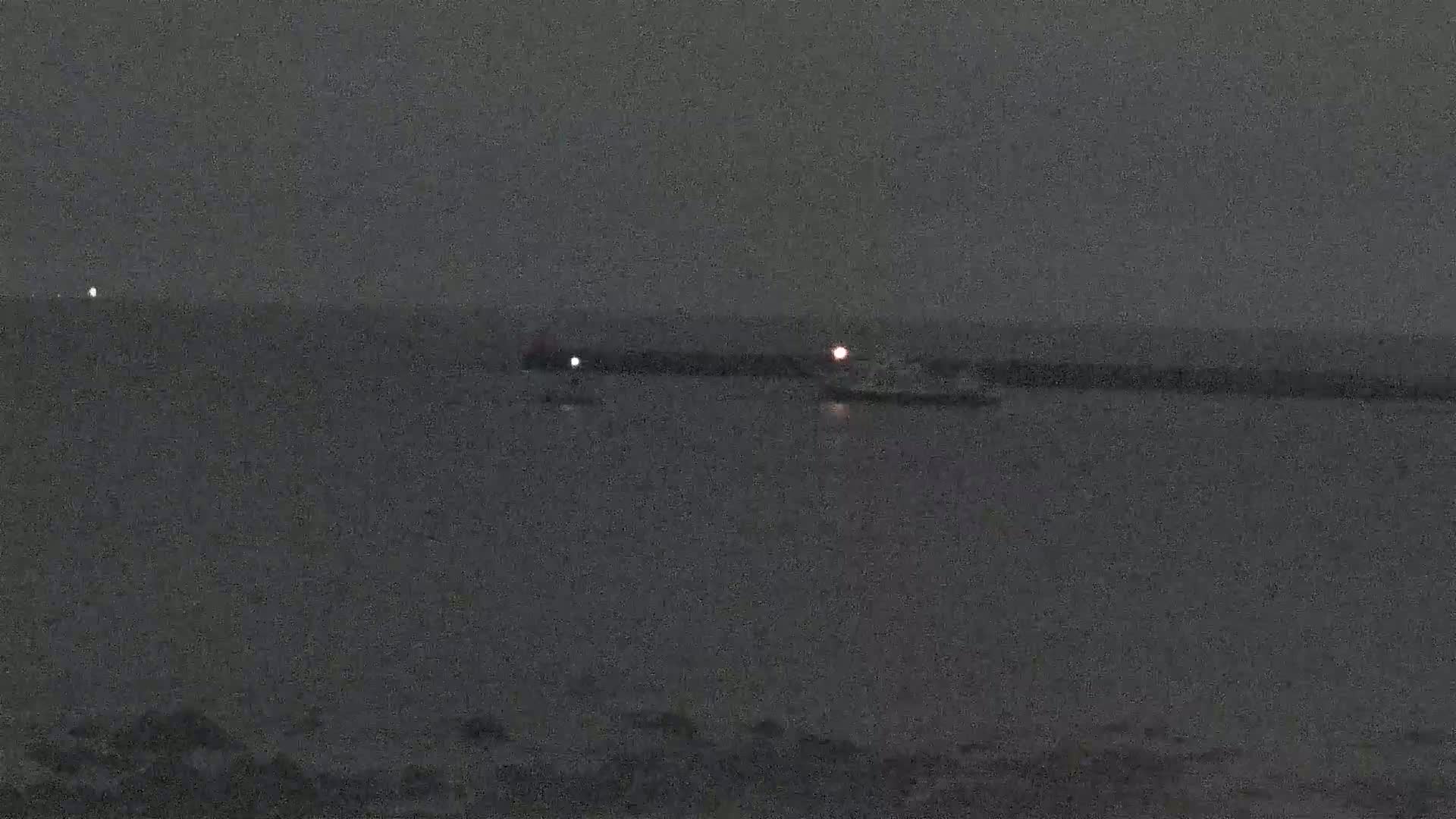 At night, a small boat with a light is near a long dark structure in the water, with another light visible in the distance.