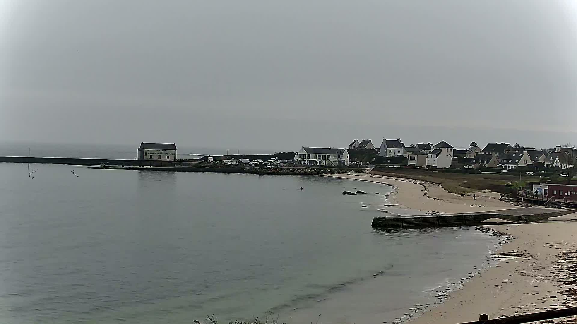 A calm body of water laps a sandy beach in front of a small coastal town under an overcast sky.