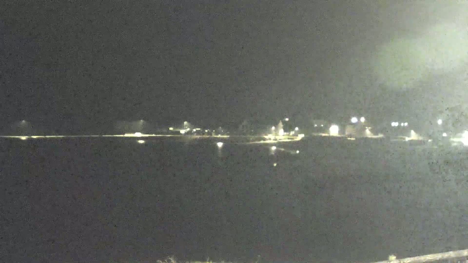 A dark, nighttime view shows a body of water reflecting the lights of a distant shoreline.
