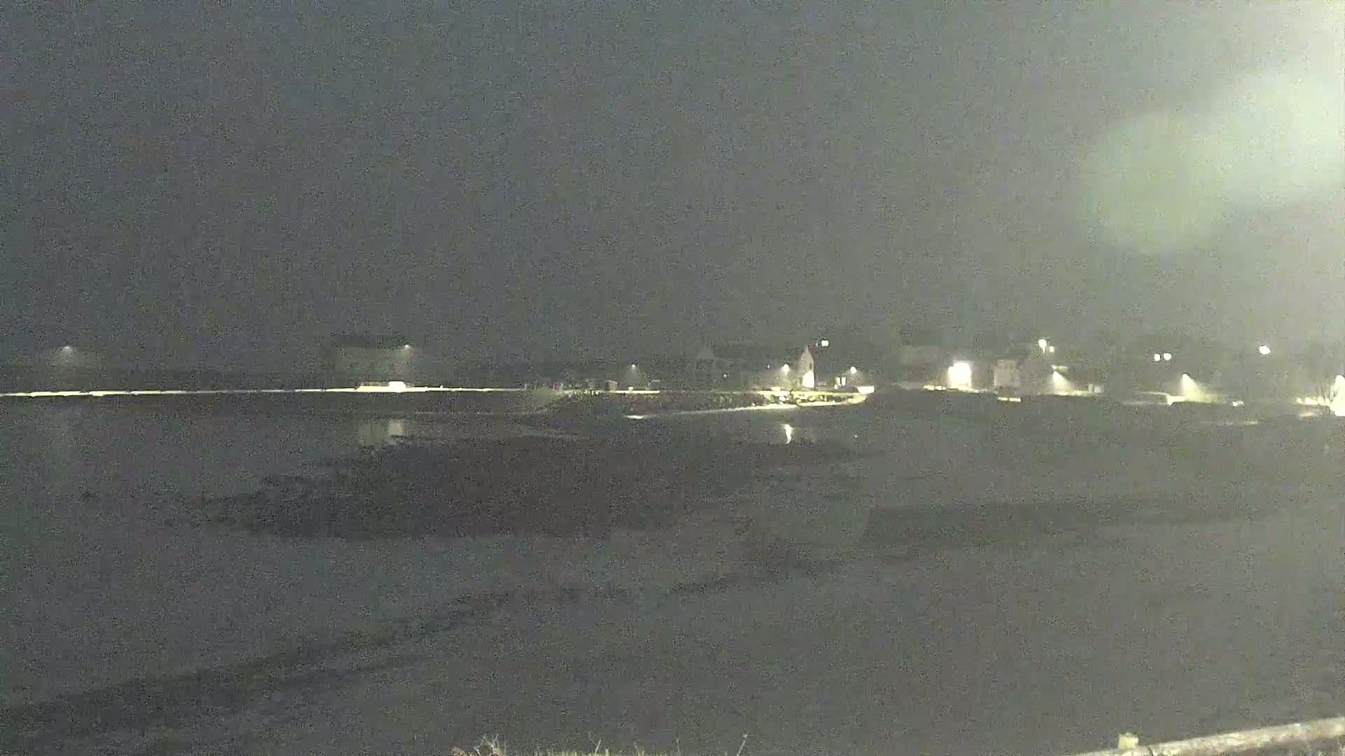 A dark, nighttime view of a calm body of water with faintly lit buildings and land in the distance.