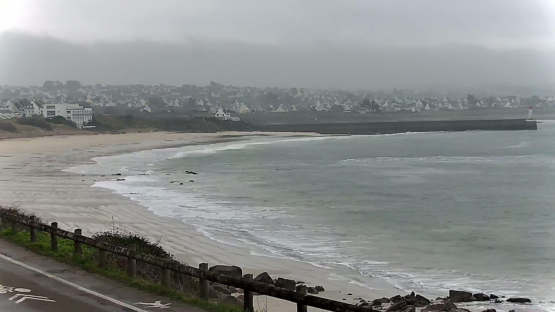 A sandy beach with gentle waves curves around a rocky point toward a distant town under an overcast sky.