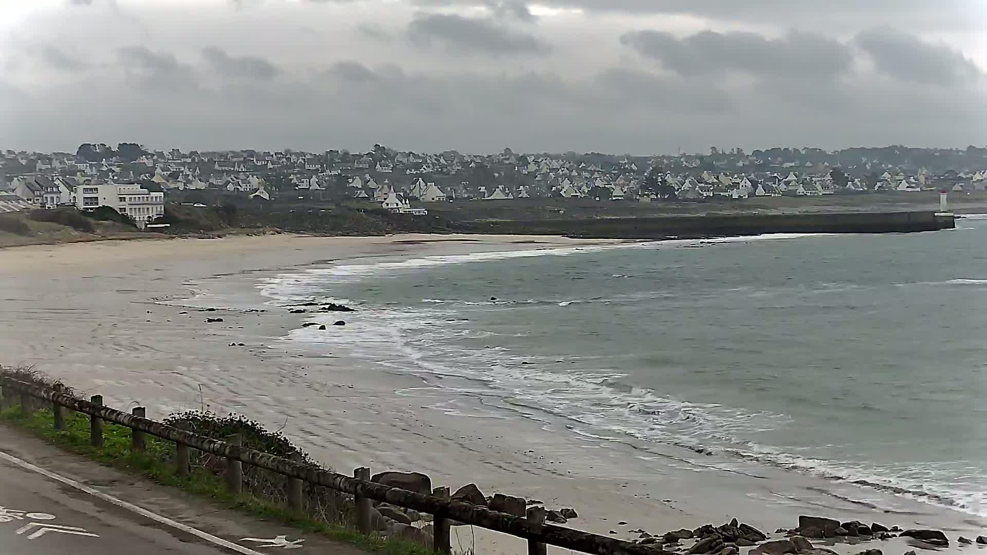 A sandy beach with gentle waves under an overcast sky overlooks a town of white buildings.