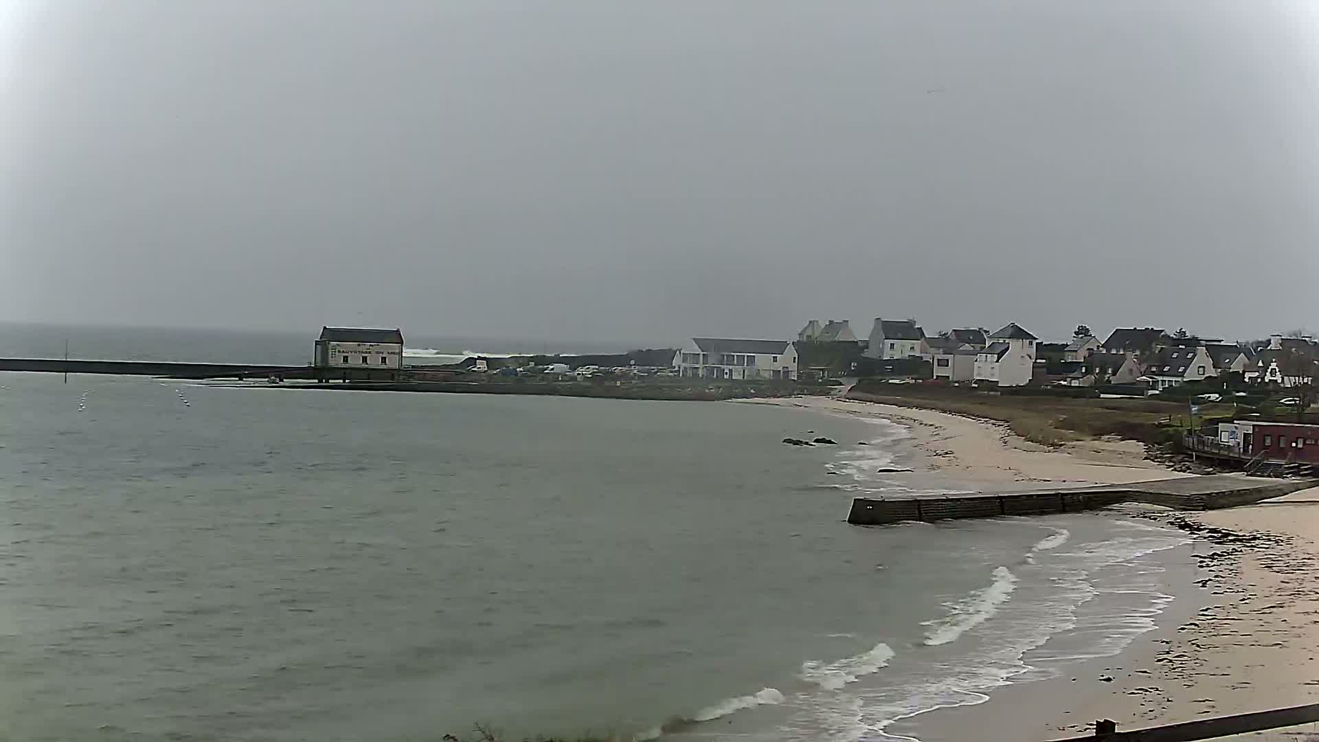 A coastal town with light-colored buildings sits beside a sandy beach and calm, gray water under an overcast sky.