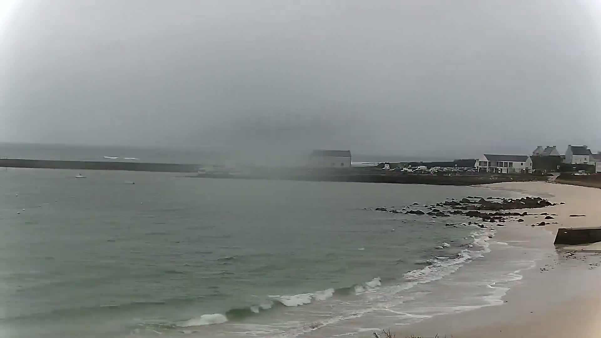 A hazy, overcast day reveals a calm sea with small waves lapping a sandy shore near a small town with several buildings.