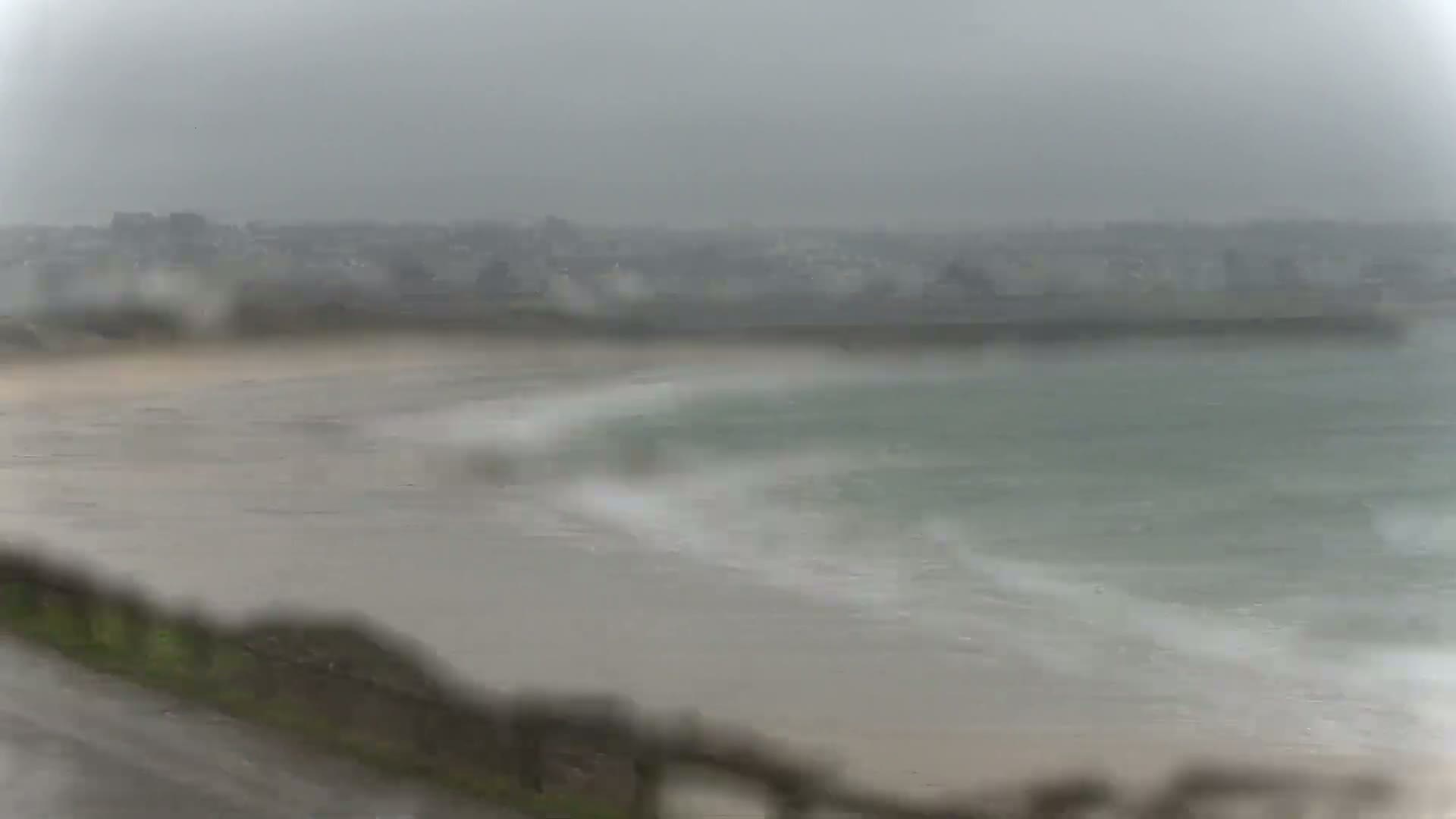 A rain-blurred view shows a beach and ocean with a town in the distance under a gray, overcast sky.