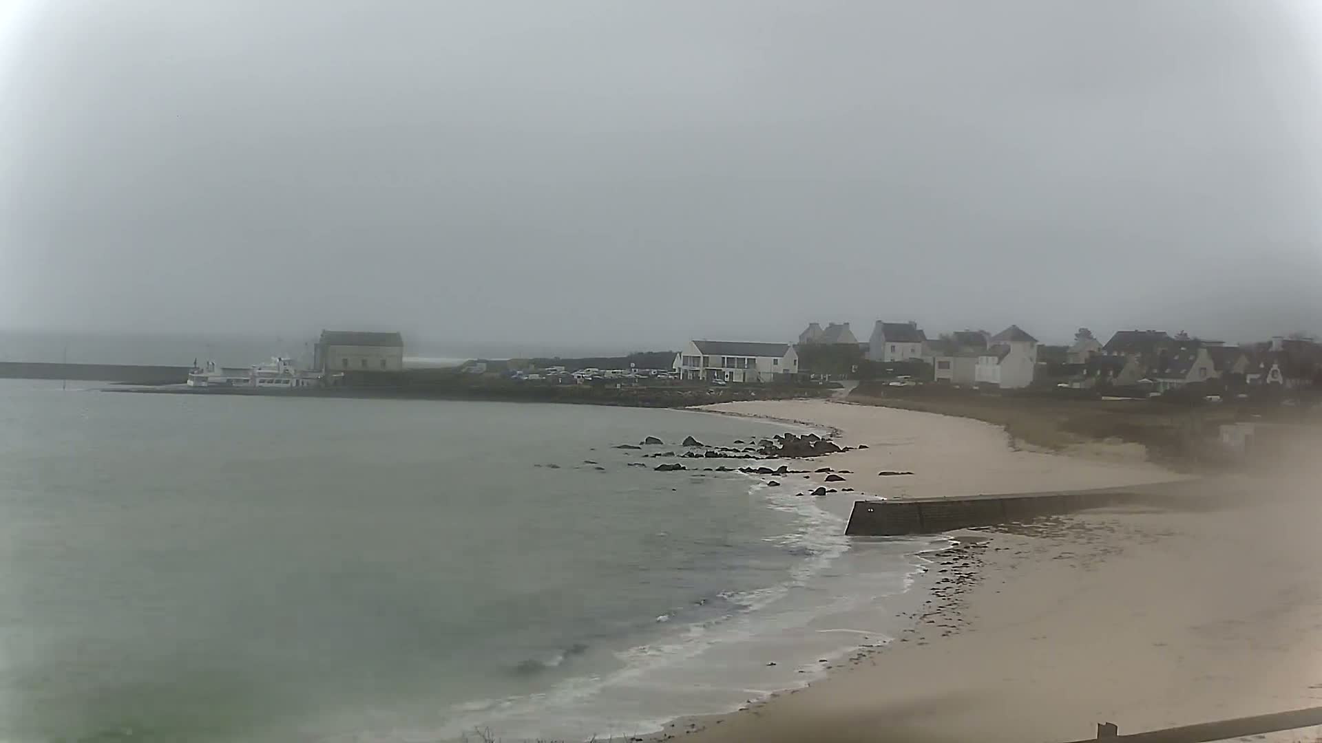 A hazy, overcast day reveals a small coastal town with a sandy beach, a low sea wall, and a boat docked near a building at the water's edge.