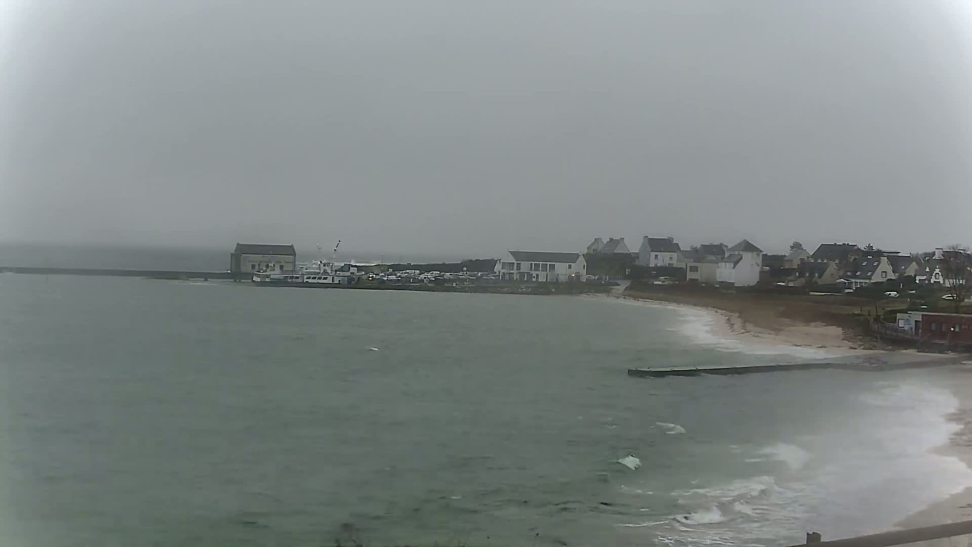 A small coastal town with a harbor and sandy beach is seen under an overcast sky, with choppy water visible in the foreground.