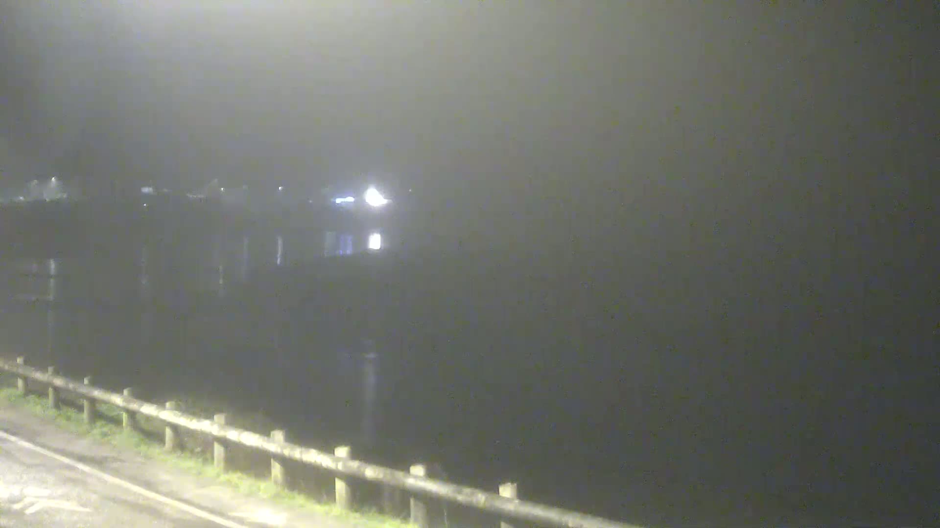 A dimly lit, foggy night scene shows a body of water with a building and a roadside barrier visible in the distance.