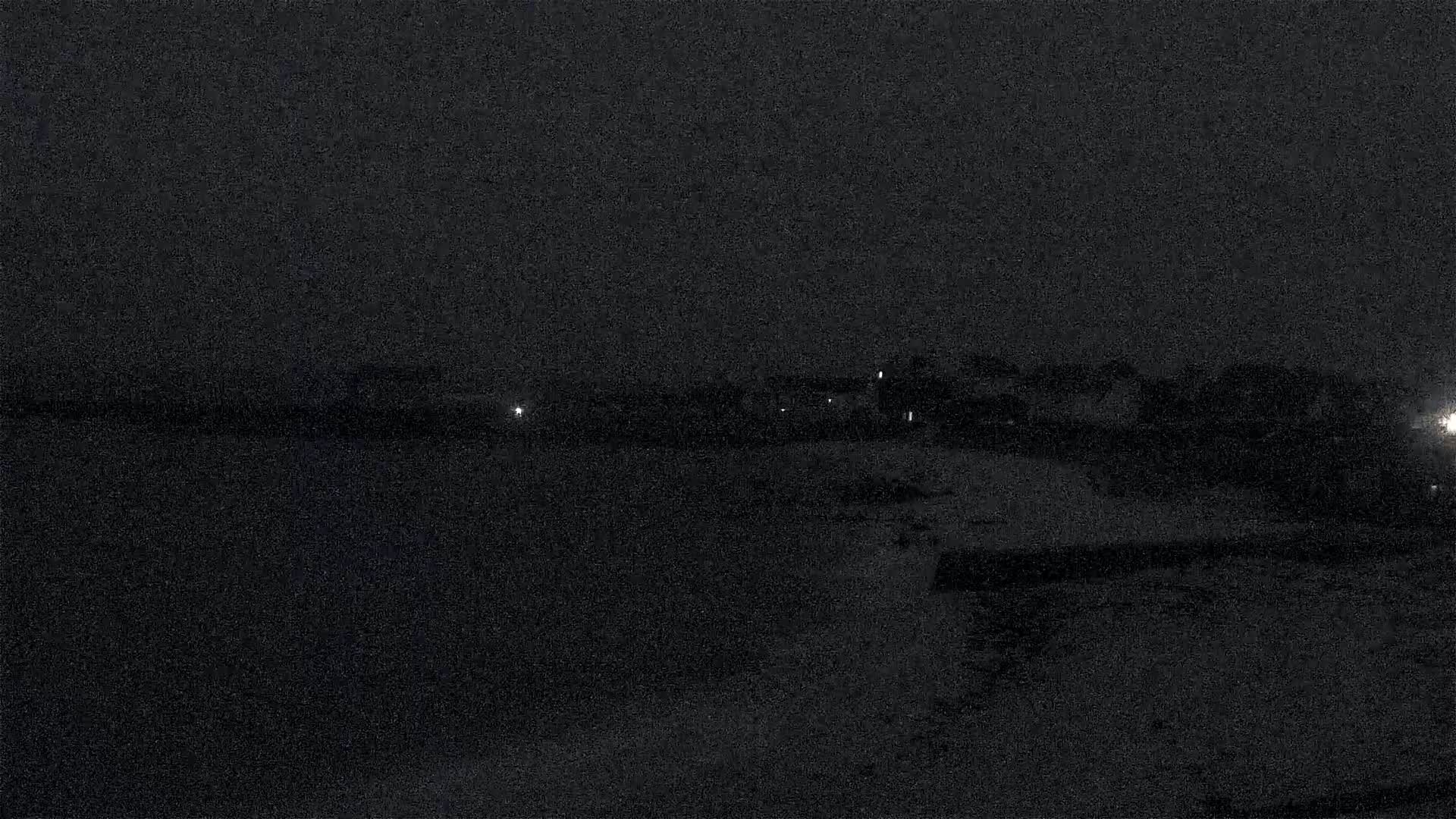A dark, nighttime scene shows a body of water in the foreground and a dimly lit shoreline with buildings in the background under a dark sky.