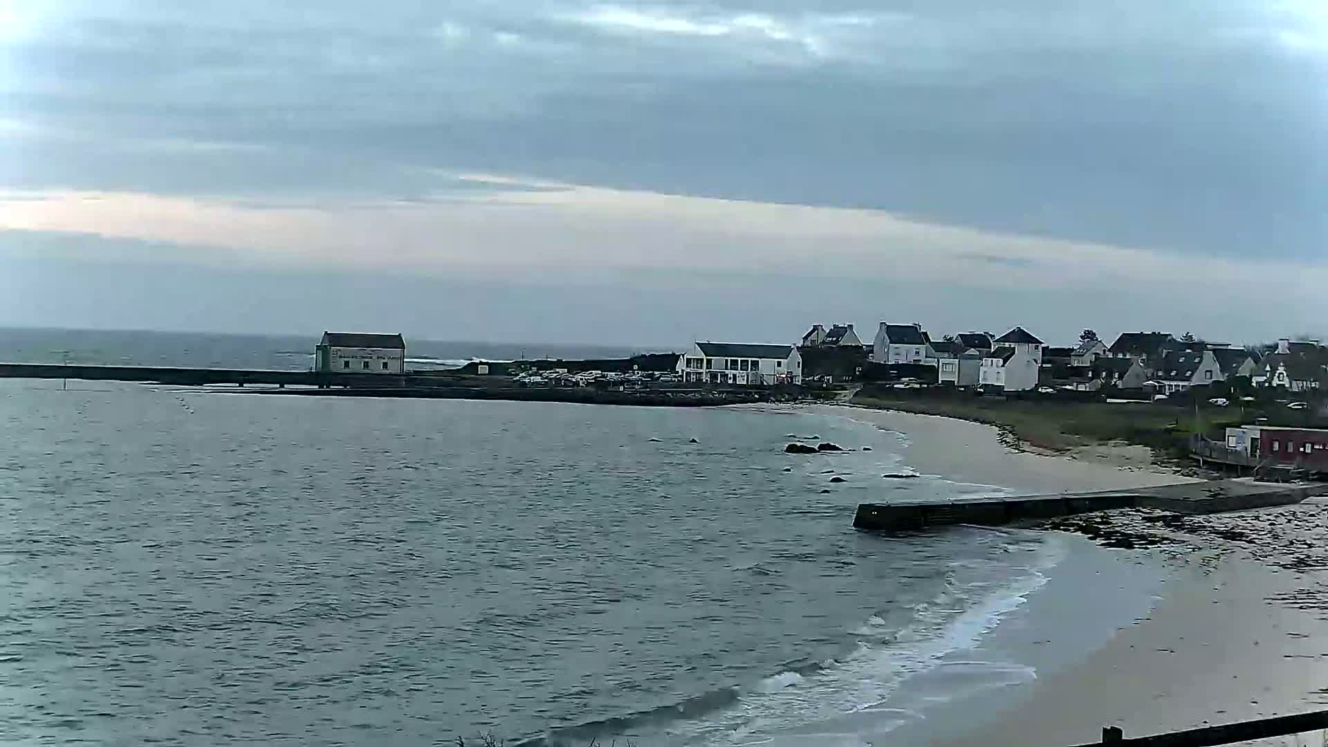 A coastal town with white buildings and a sandy beach is visible under a cloudy sky, with gentle waves lapping the shore.