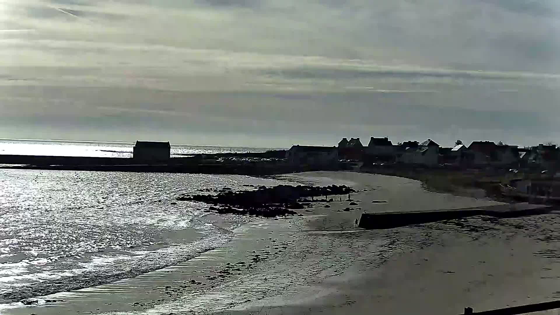 A mostly cloudy day shows a calm sea lapping a sandy beach near a small village of houses.