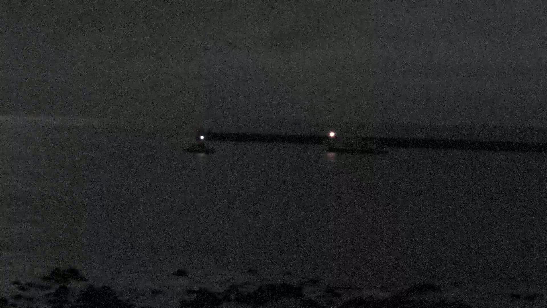 At night, two small lights are visible on the water, seemingly emanating from objects near a dark, long structure.