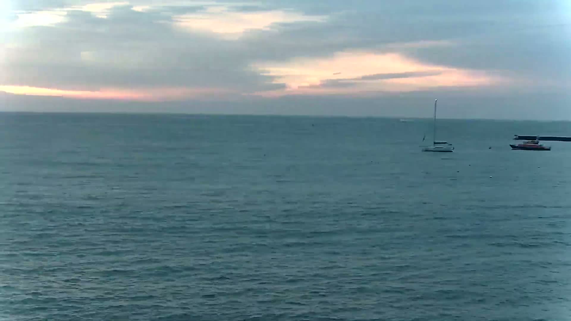 A sailboat and a small motorboat are visible on a calm, gray-blue sea under a mostly cloudy sky with a hint of pink at the horizon.