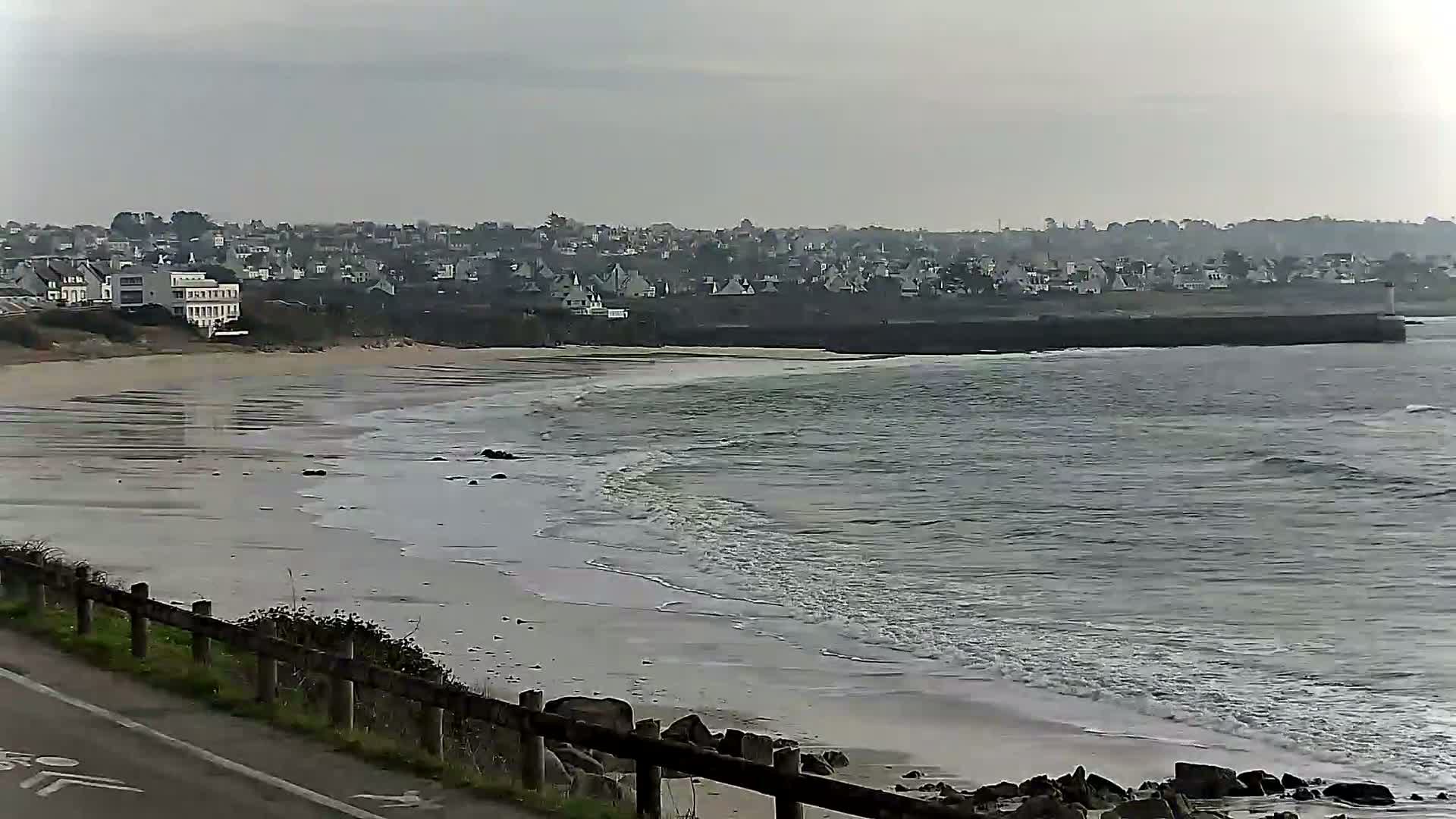 A sandy beach with gentle waves laps the shore under an overcast sky, with a line of houses visible in the distance.
