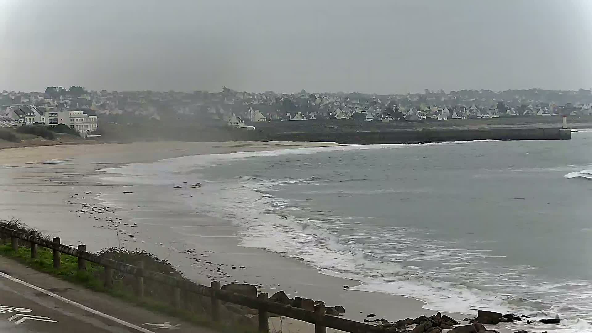 A sandy beach with gentle waves rolls onto the shore under an overcast sky, with a town and buildings visible in the background.