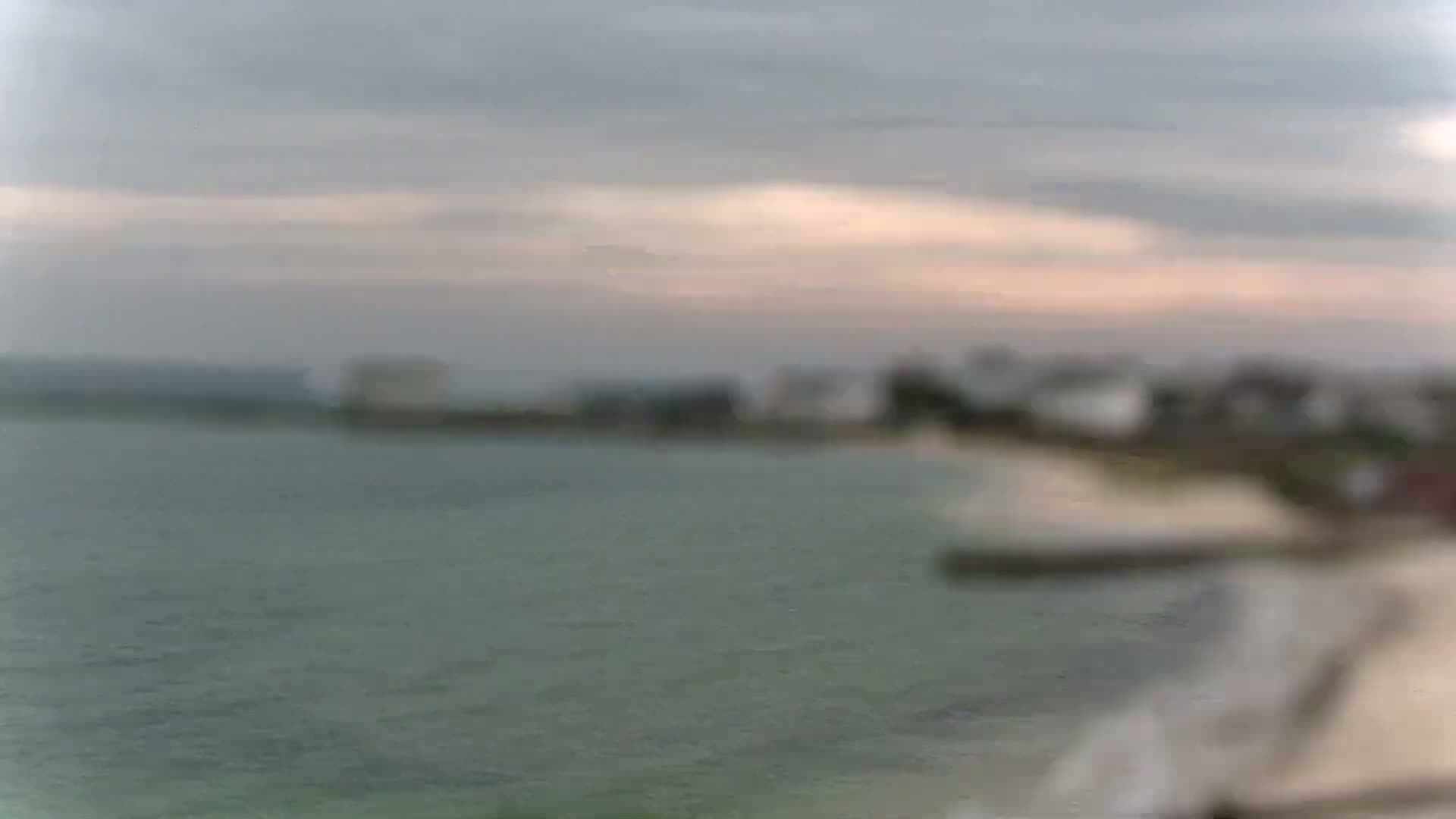 A blurry image shows a calm body of water under an overcast sky with a faint pinkish hue near the horizon, and a distant shoreline with buildings.