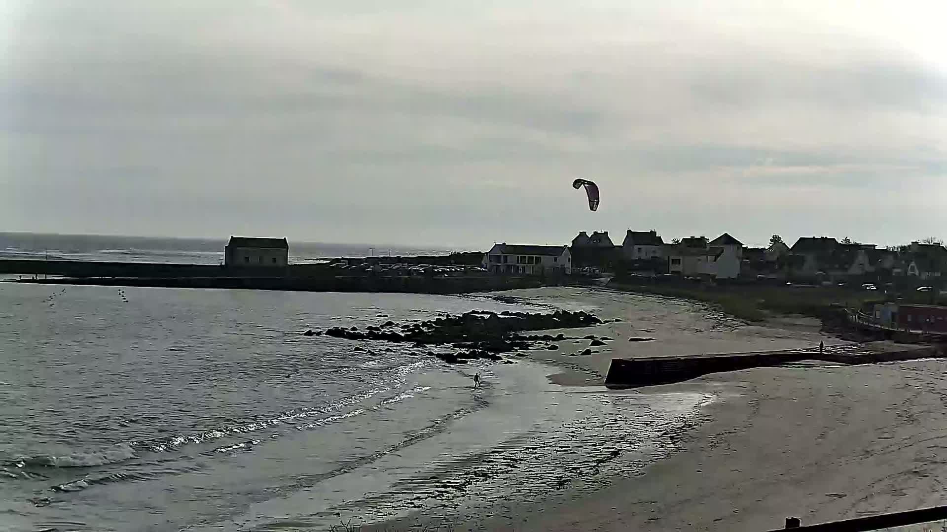 A kite surfs above a calm, grey sea lapping a sandy beach near a small town under an overcast sky.