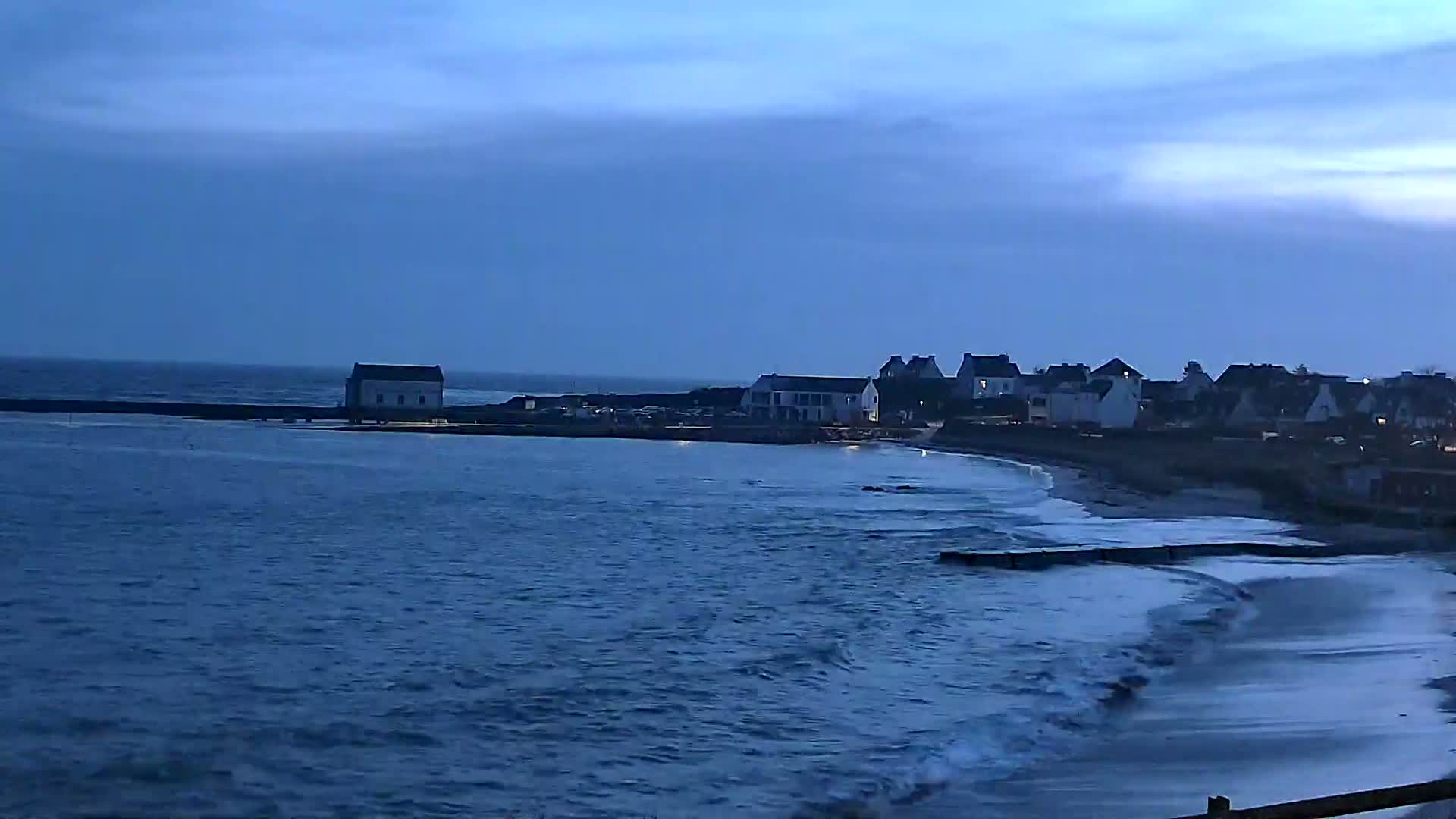 A coastal town is visible at dusk, with small waves breaking on a sandy beach under a mostly clear, dark blue sky.