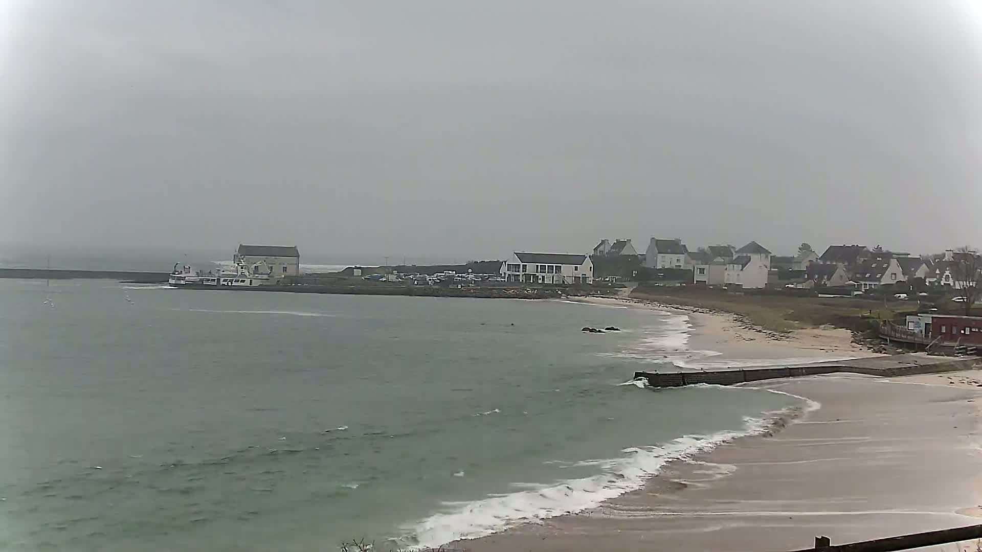 A small coastal town is visible on a grey, overcast day, with a boat docked near a building and a sandy beach with gentle waves lapping the shore.