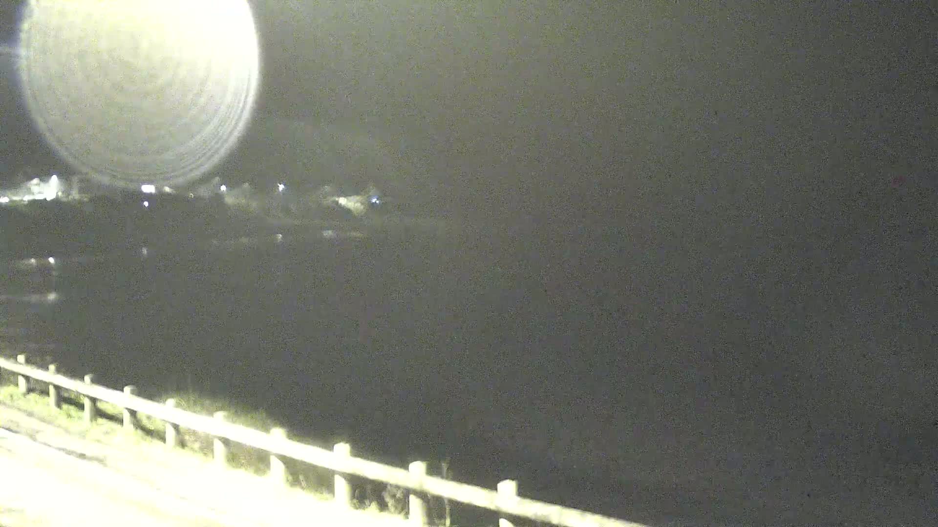 A nighttime view shows a dark body of water, a dimly lit shoreline with a fence, and a bright light source creating a glare in the upper left corner.