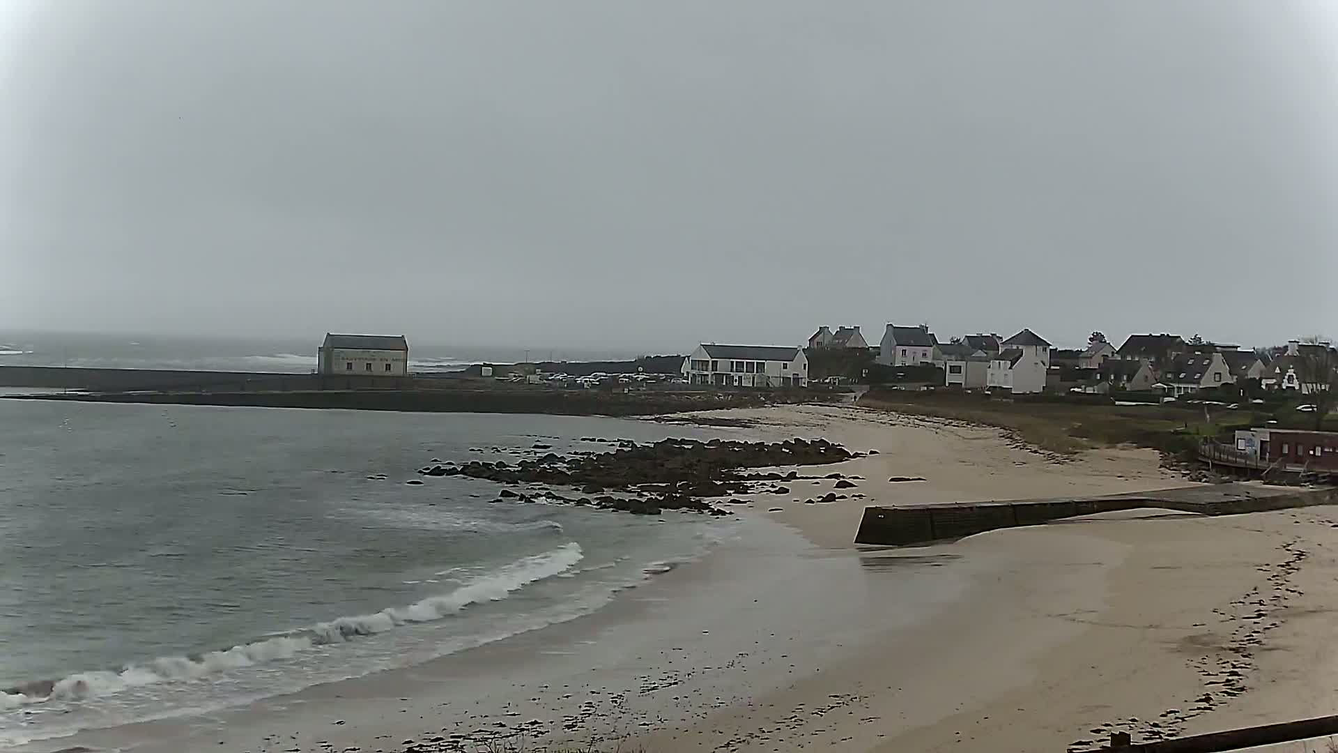 A sandy beach with small waves laps against a rocky shoreline, behind which sits a small village under an overcast sky.